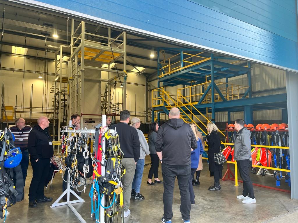 A group of people gathered in a warehouse with safety equipment and harnesses on display racks.