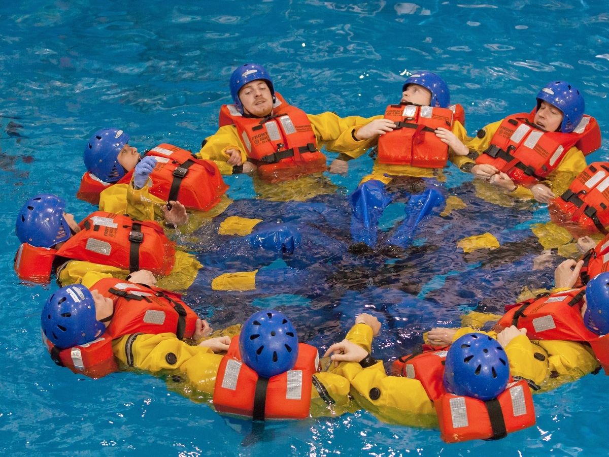 A group of people wearing orange life jackets and helmets form a circle while floating in a pool, participating in a training exercise.