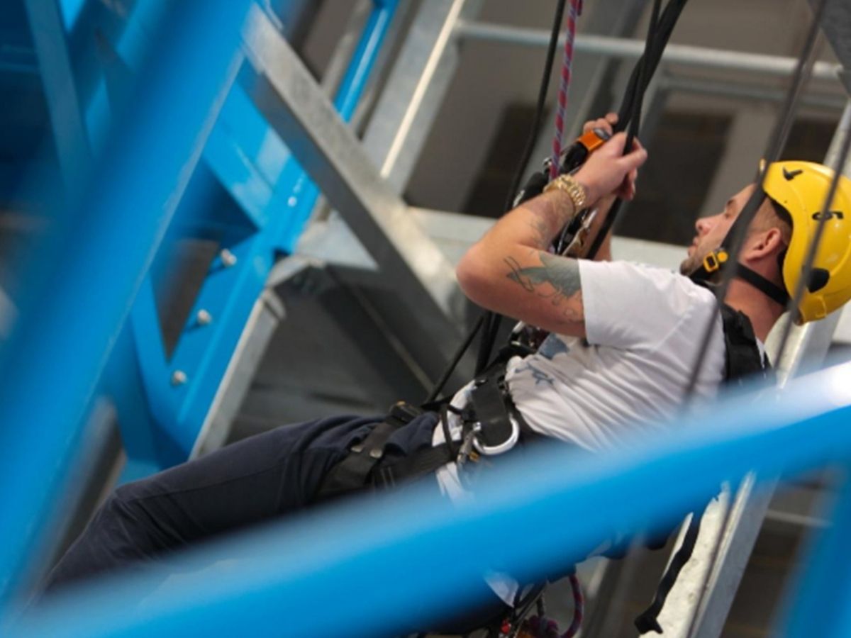 A man wearing a harness and yellow helmet is hanging from ropes doing rope access training.