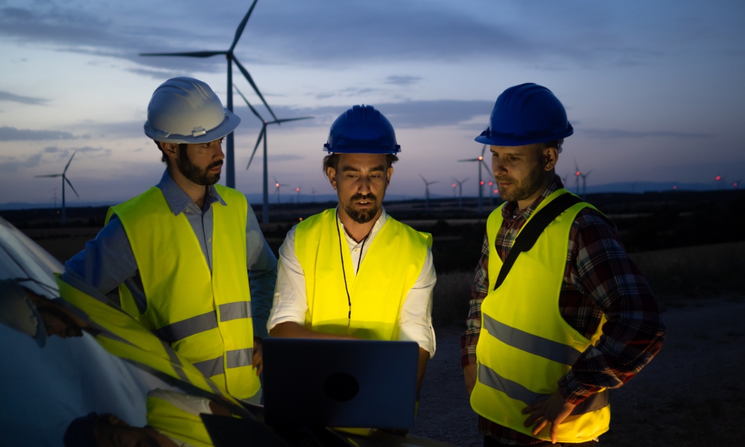 Three workers in safety gear and helmets undergo training as they examine a laptop near wind turbines at dusk.