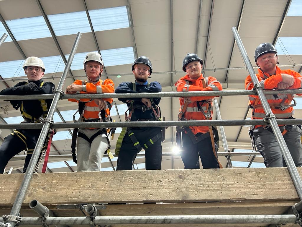 scaffolding apprentices in hard hats and harnesses looking down from scaffolding platform