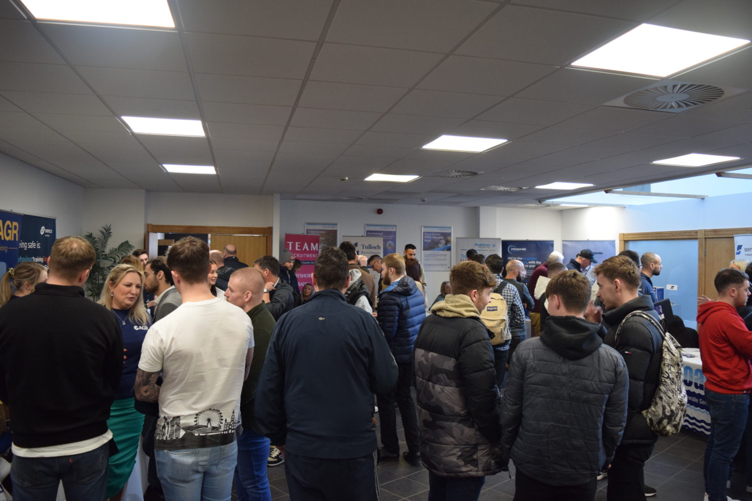 A group of people mingling in a conference room, engaging in conversations near informational booths and banners promoting upcoming training sessions.
