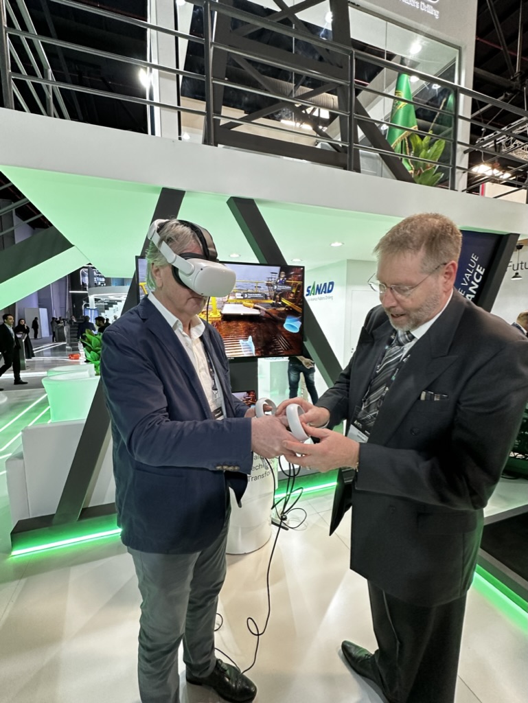 Two men at an exhibition; one wearing a VR headset and holding a controller, immersed in a training simulation, while the other assists. The booth is bathed in green lighting with a screen displaying dynamic visuals in the background.