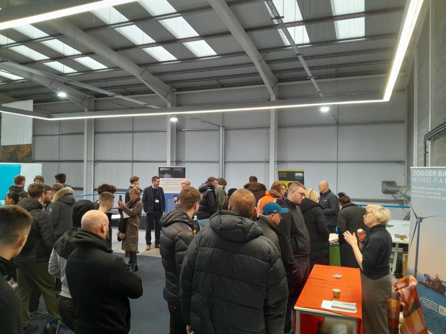 A group of people gathered indoors for a training event, near display tables and banners. Many are wearing jackets. The space has high ceilings and overhead lighting, creating an inviting atmosphere for learning and networking.