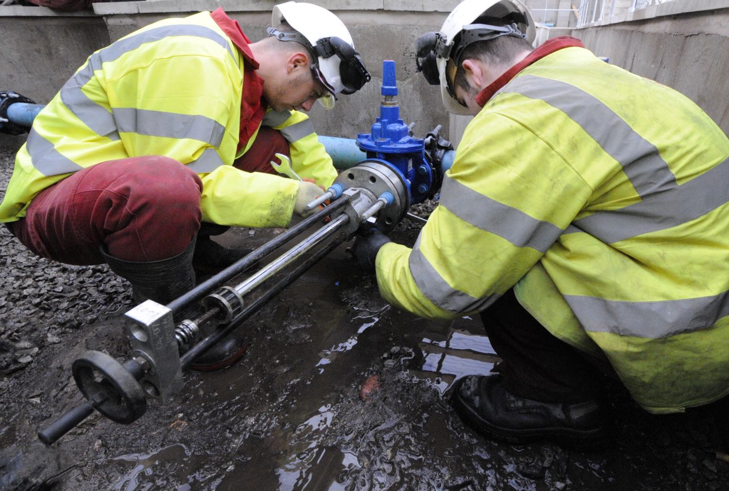 Two workers in safety gear, utilizing their training, repair a pipe outdoors, surrounded by wet ground.