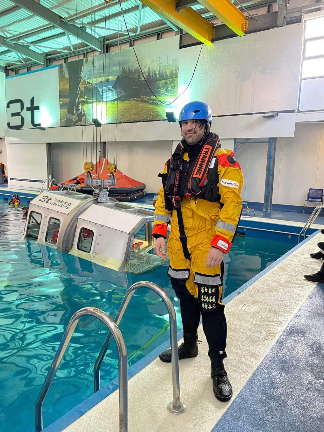 A person in water survival gear stands by a training pool, where a mock helicopter is partially submerged for a realistic training scenario.