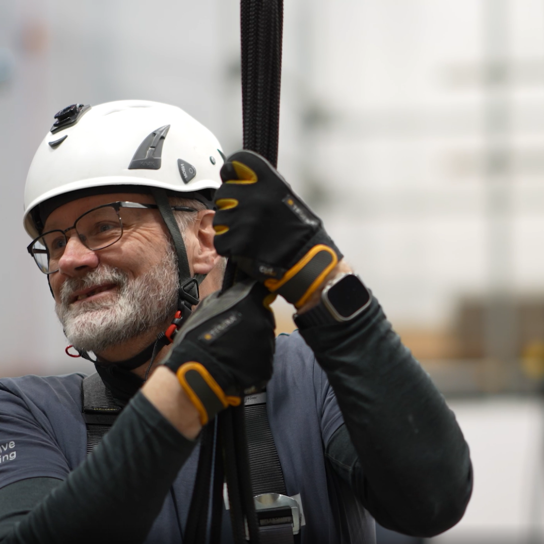 A man in a helmet and gloves holds a safety rope, wearing a dark shirt and glasses during training.
