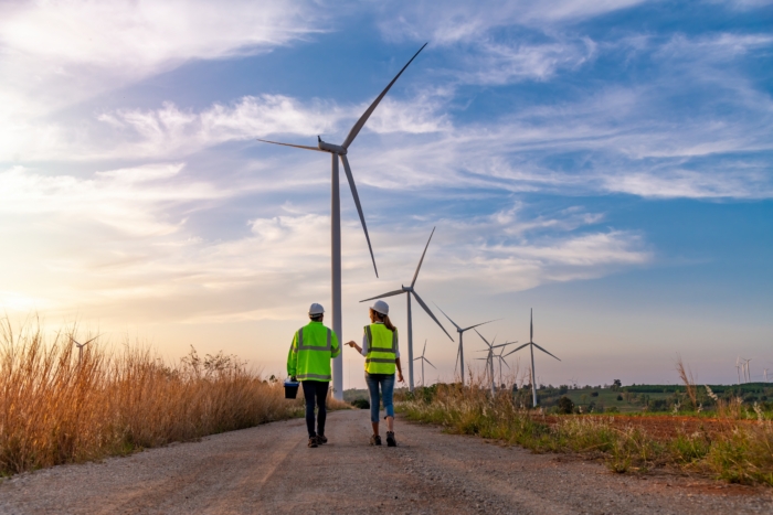 Two people in safety vests walk on a path surrounded by wind turbines under a blue sky, participating in valuable training on renewable energy.