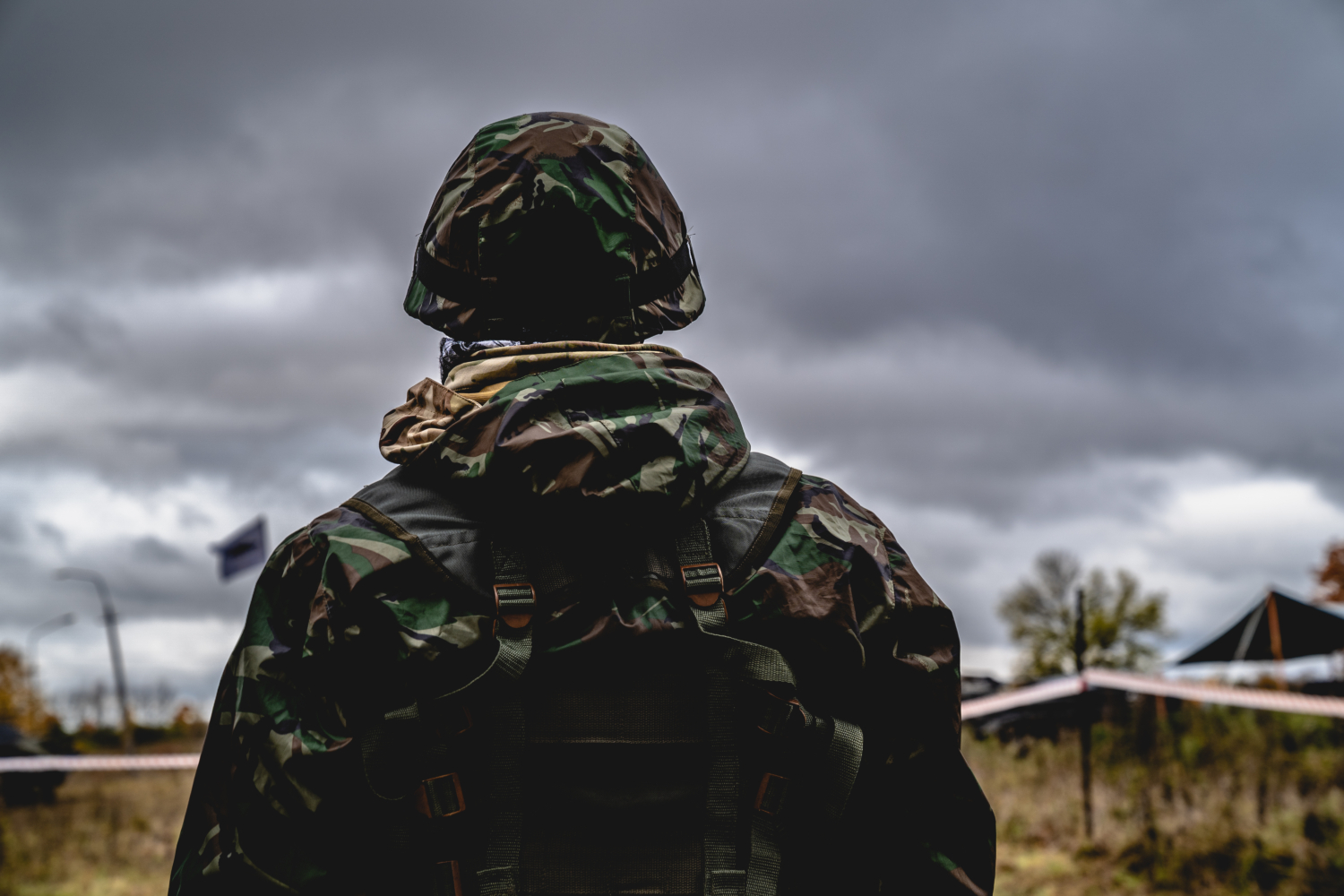 A person in camouflage military gear stands outdoors, possibly engaging in training exercises, under a cloudy sky.