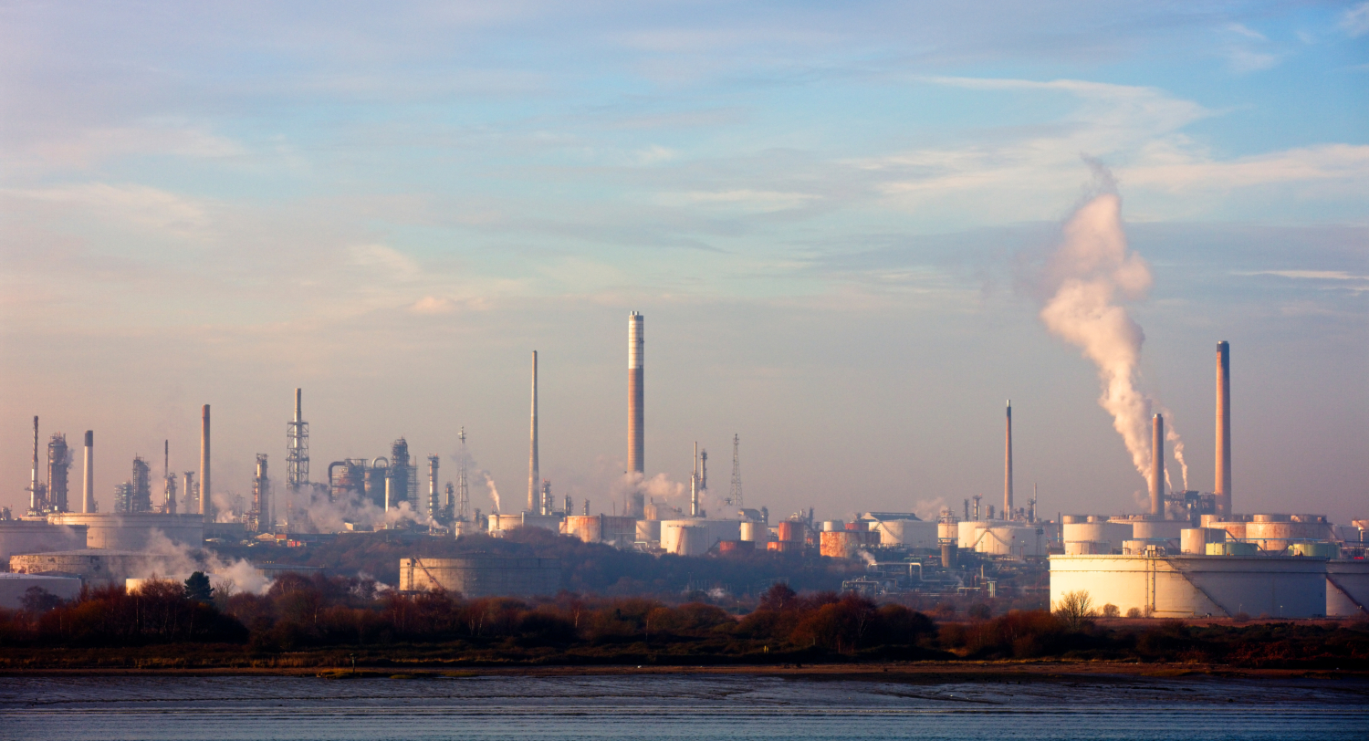 An industrial refinery landscape sprawls beneath a blue sky with scattered clouds, its chimneys puffing smoke like sentinels in training, synchronized and steadfast.