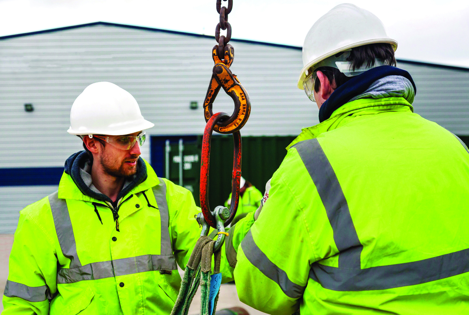 Two construction workers in high-visibility jackets and helmets are skillfully handling a large hook and chain outdoors near a building, showcasing their training.