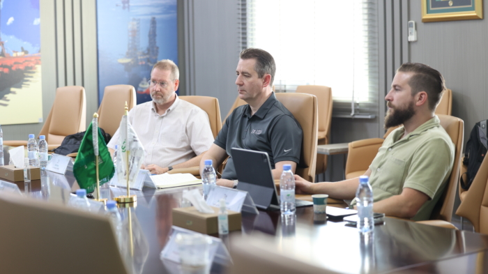 Four men seated at a conference table engage in a training meeting. Various items, including laptops and water bottles, are visible on the table. Flags are also present.