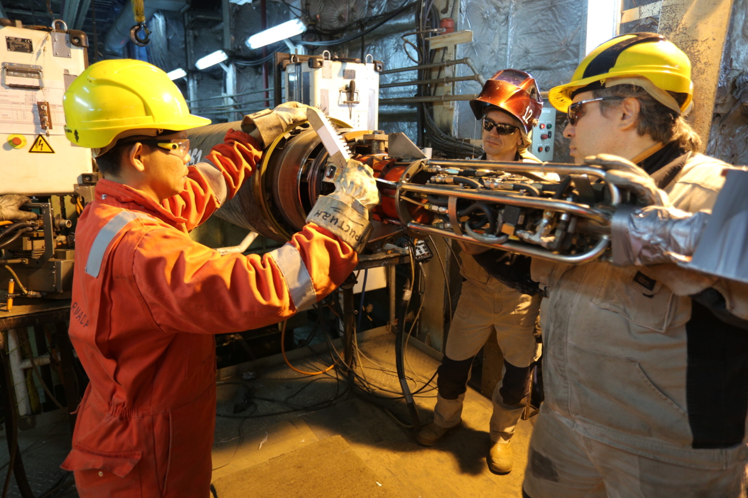 Three workers in protective gear are participating in training, collaborating in an industrial setting while handling machinery parts with tools and equipment around.