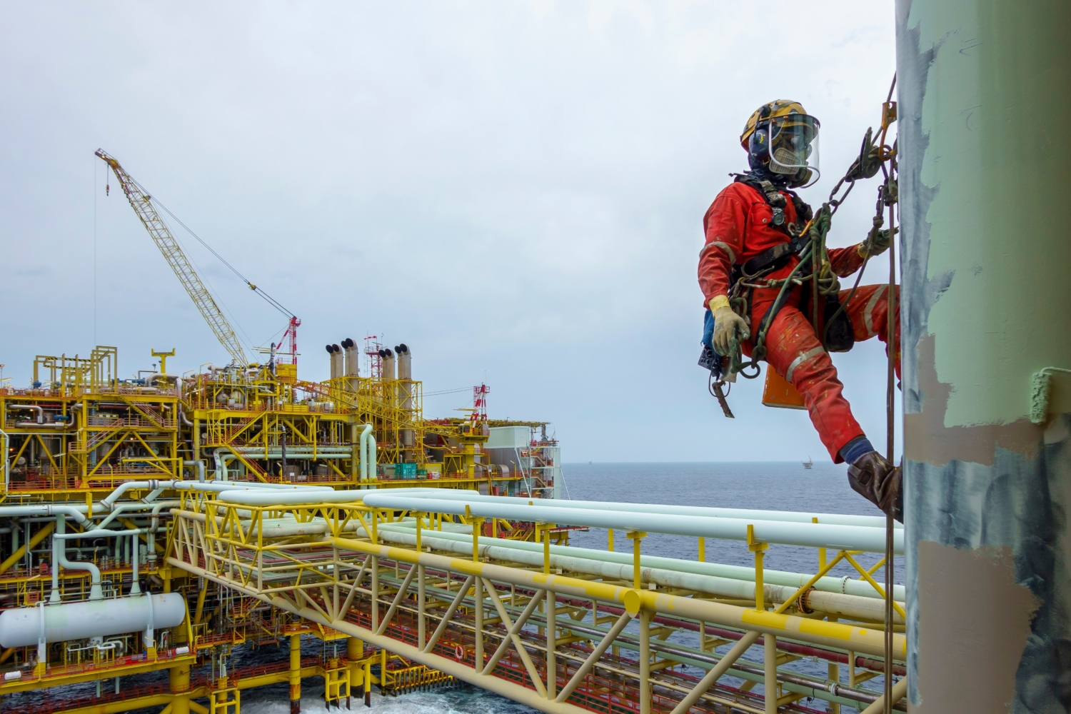 A worker in safety gear, demonstrating expert training, is suspended on ropes inspecting a vertical structure on an offshore oil rig, with ocean and platform machinery visible in the background.