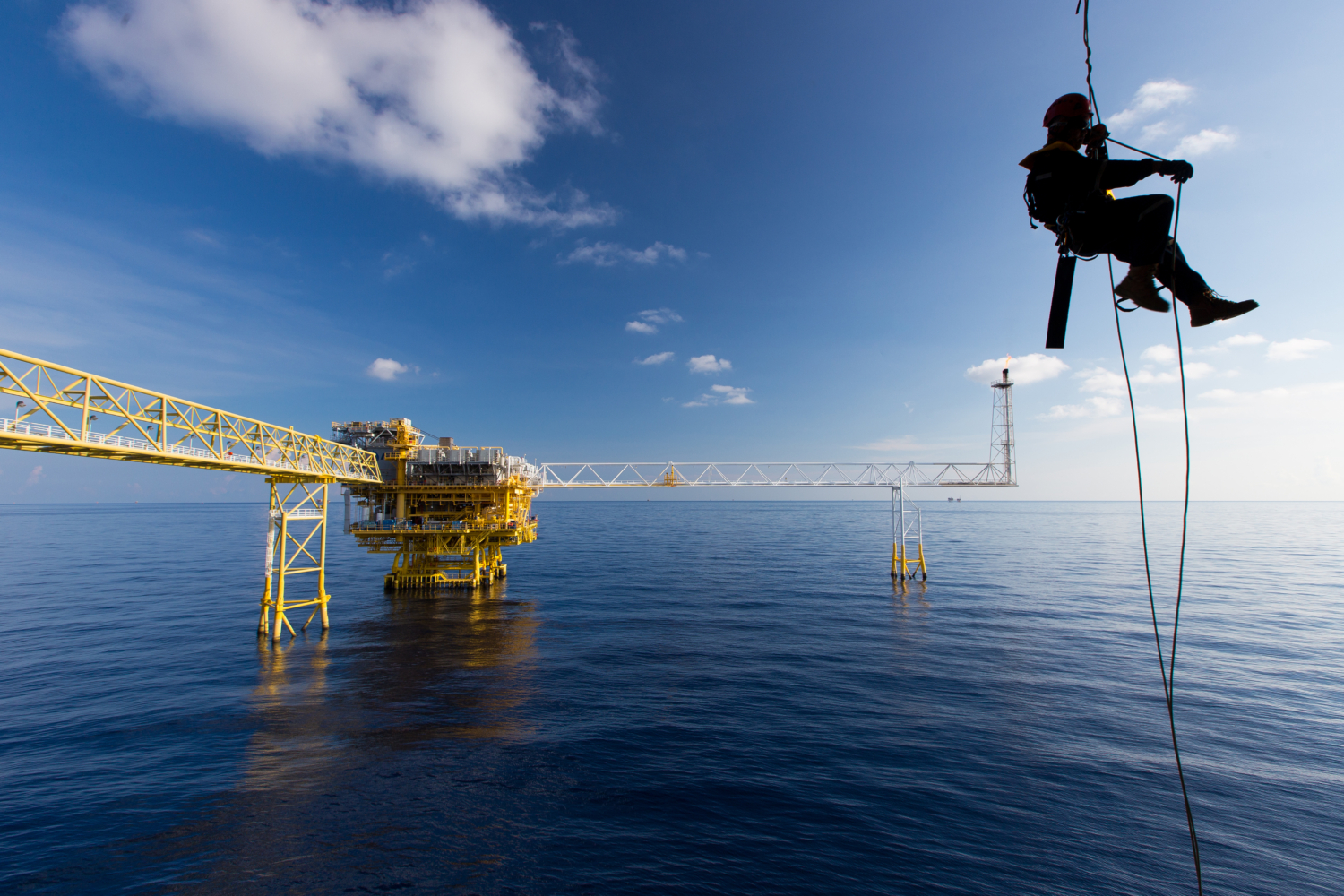 A worker, undergoing training, is suspended by a safety rope above the ocean, with an offshore oil platform in the background under a blue sky with scattered clouds.