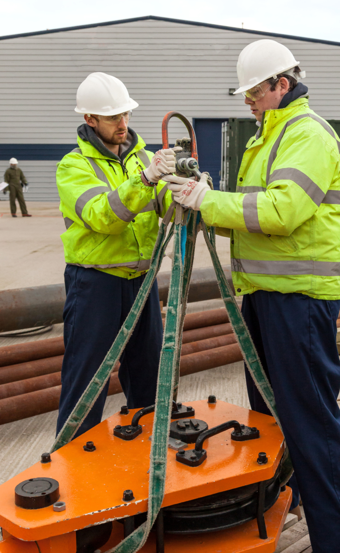 Two workers in high-visibility jackets and helmets adjust a heavy-duty lifting clamp with straps outside a warehouse, honing their skills through practical training.