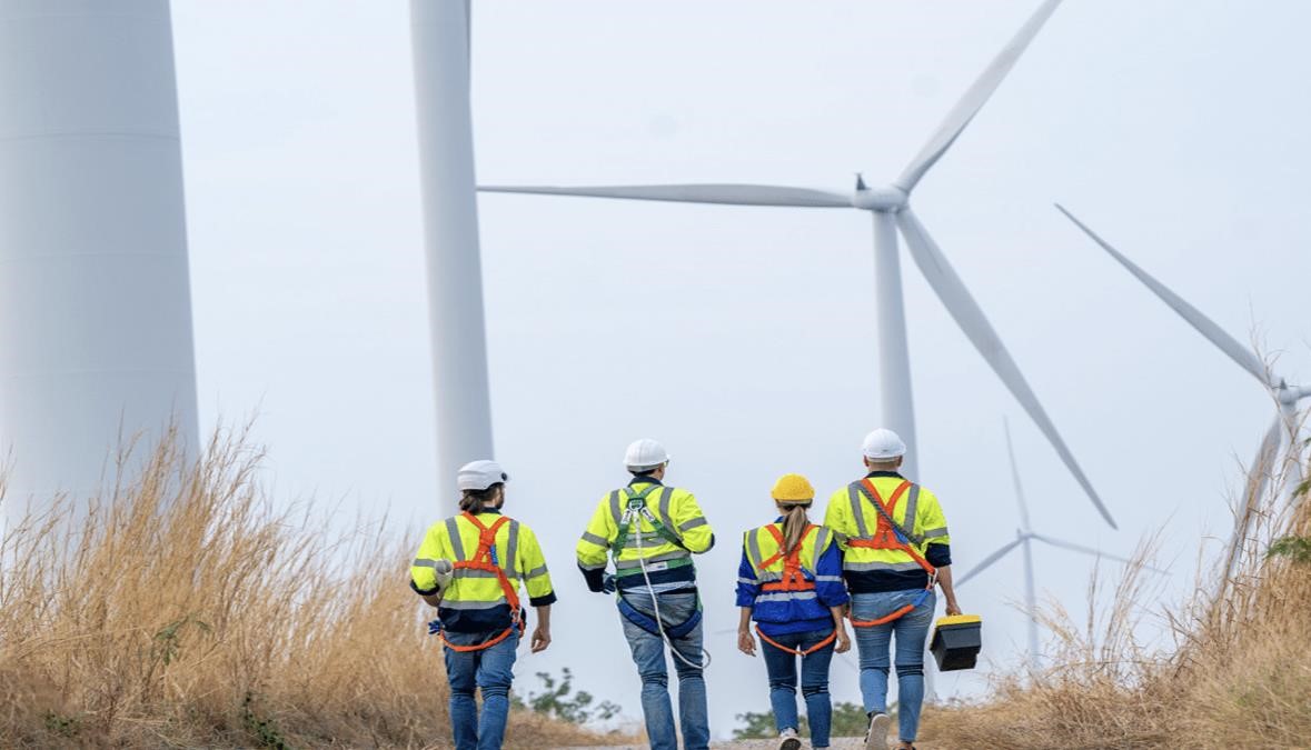 Four workers in safety gear, fresh from a training session, walk toward wind turbines in a field.