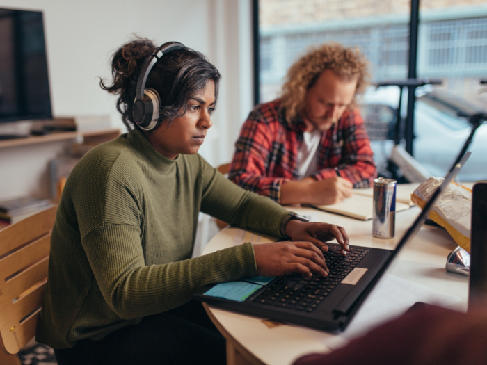A person with headphones works on a laptop at a table, focused on training modules, while another person writes in the background.