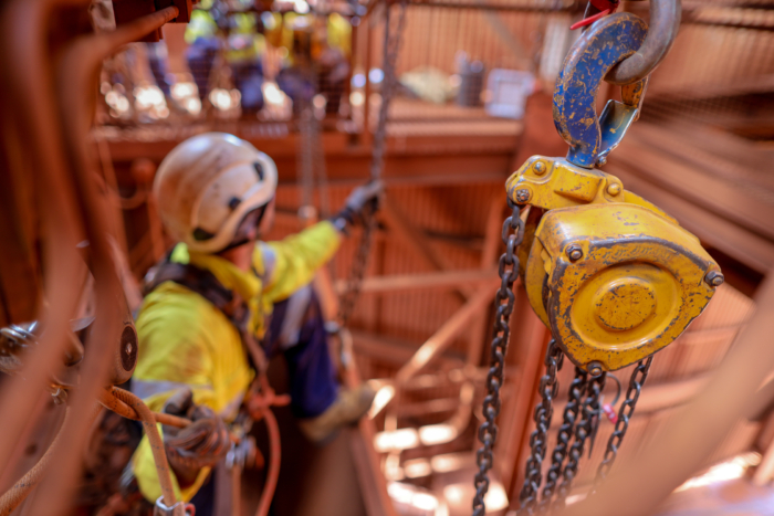A construction worker in safety gear, undergoing training, is suspended by ropes next to a yellow hoist with chains.