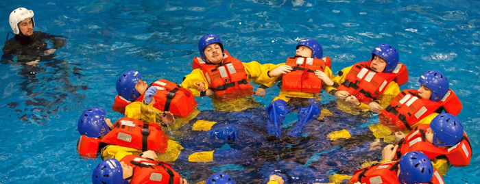 People in yellow suits and helmets float in a pool in a circular formation, wearing orange life vests as part of their training. A person in black stands nearby.