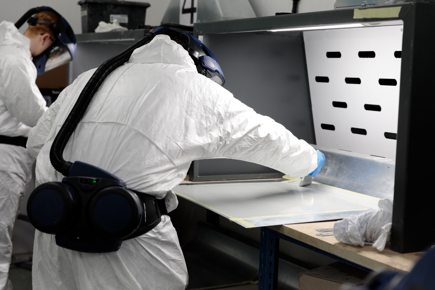 A person in protective gear is engaged in training at a ventilated table, meticulously applying a substance to a surface. Another individual, also in similar gear, observes from the background, ensuring precision and safety.