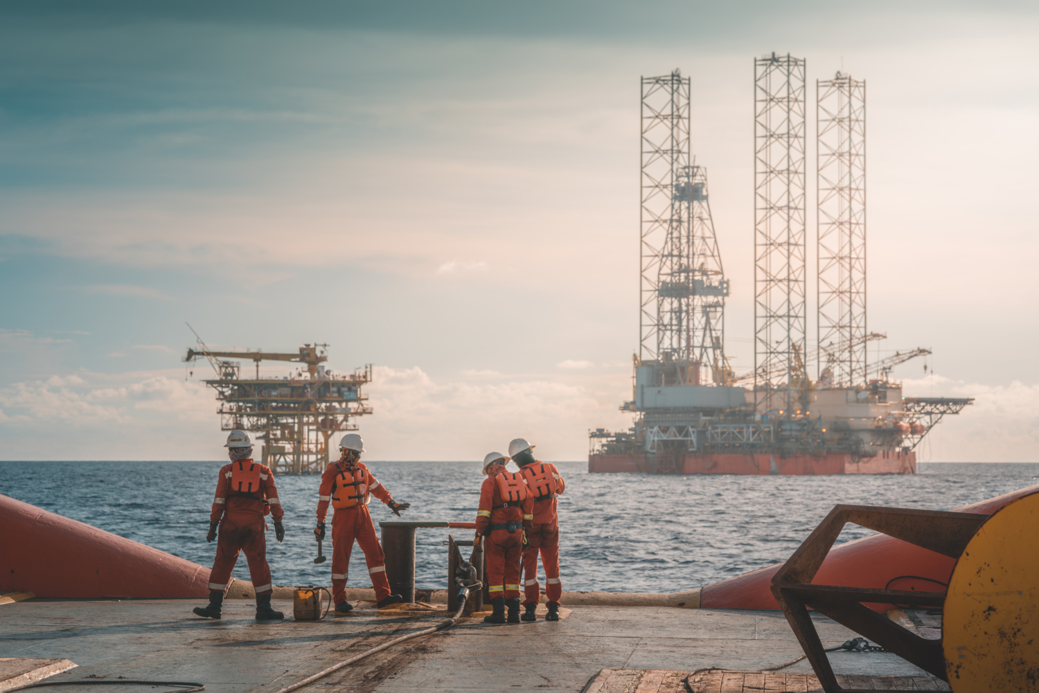 Four workers in safety gear stand on an offshore platform, honing their skills through hands-on training as they perform tasks, with two oil rigs visible in the background over the sea.
