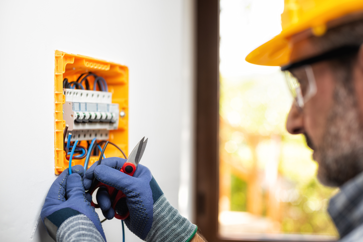 Electrician in a yellow hard hat and gloves installs a circuit breaker in a wall-mounted electrical panel.