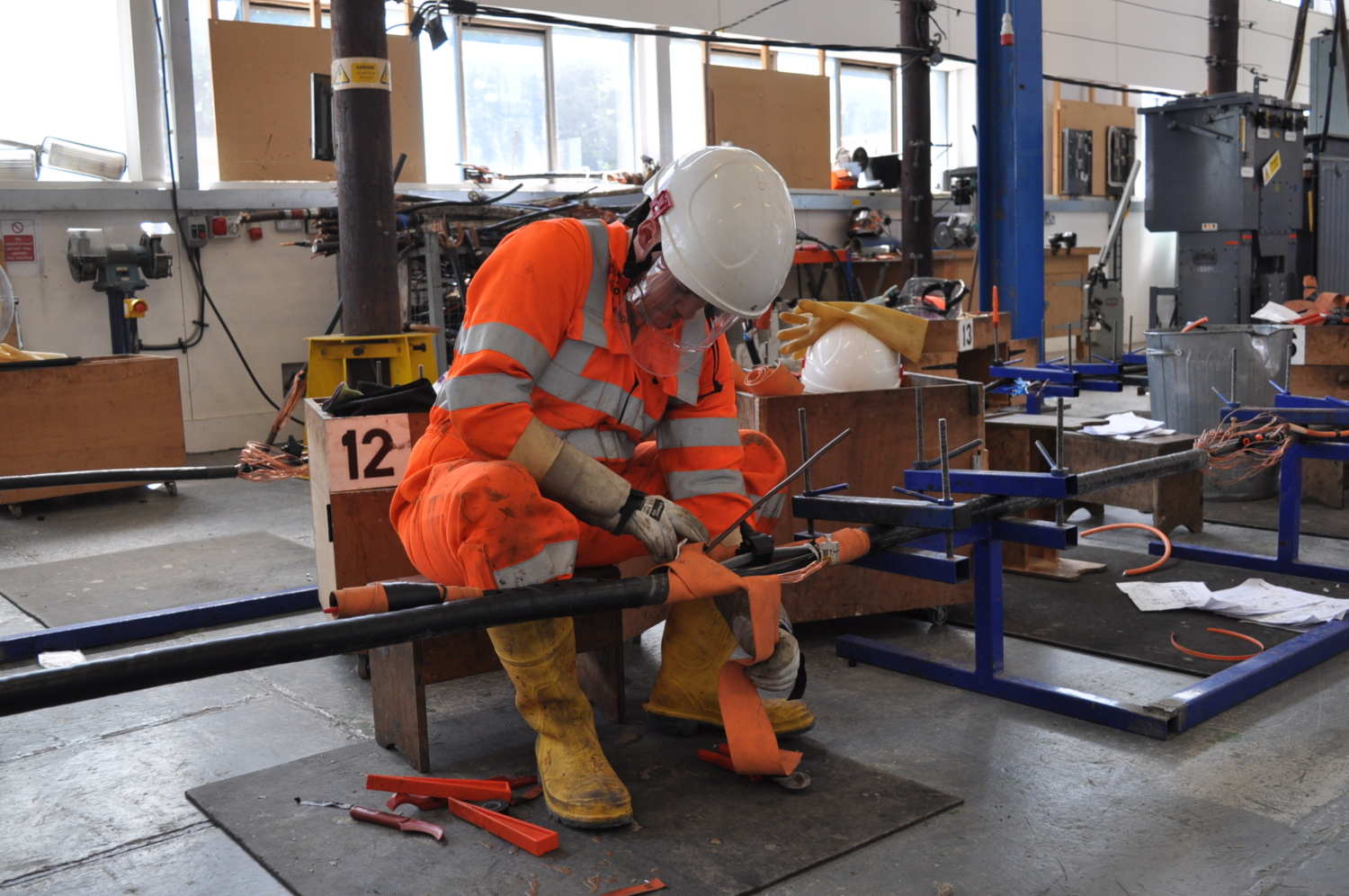 A worker in orange protective gear and a white helmet repairs equipment in a workshop, surrounded by tools and supplies.
