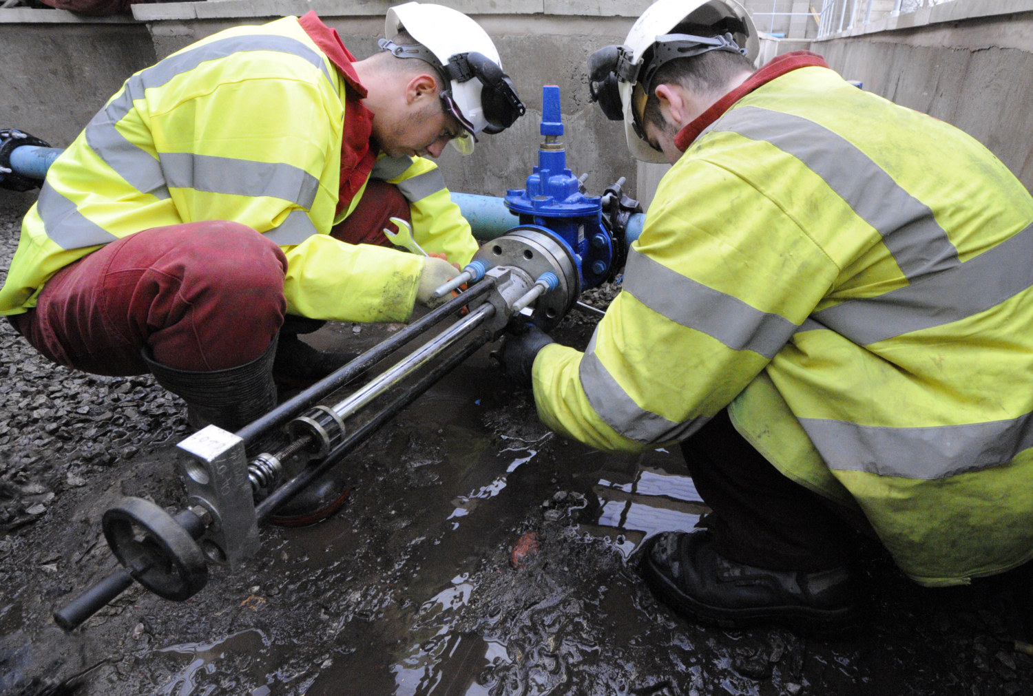 Two workers in high-visibility jackets and helmets are training while inspecting or repairing an industrial pipe outdoors, surrounded by muddy ground.