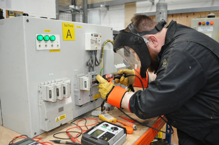 A person wearing protective gear engages in training, working on an electrical test panel with tools and meters spread across a wooden table.