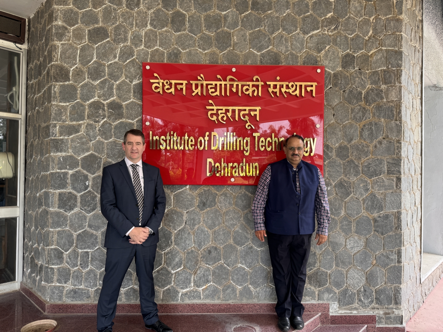 Two individuals stand in front of a red sign with text in Hindi and English that reads 