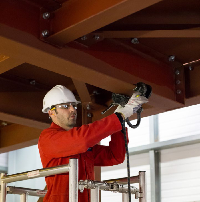 A worker in a red jumpsuit and white helmet uses a power tool on a steel beam, standing on scaffolding indoors.