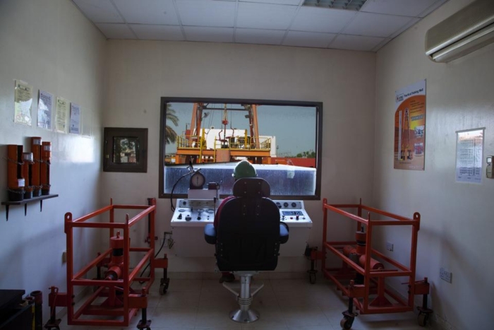 Interior of a control room with a person seated at a console, monitoring a crane lifting a container outside. Red equipment frames flank the room, and informational posters are on the walls.