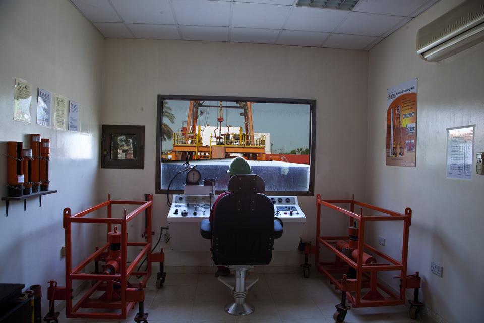 Interior of a control room with a person seated at a console, monitoring a crane lifting a container outside. Red equipment frames flank the room, and informational posters are on the walls.