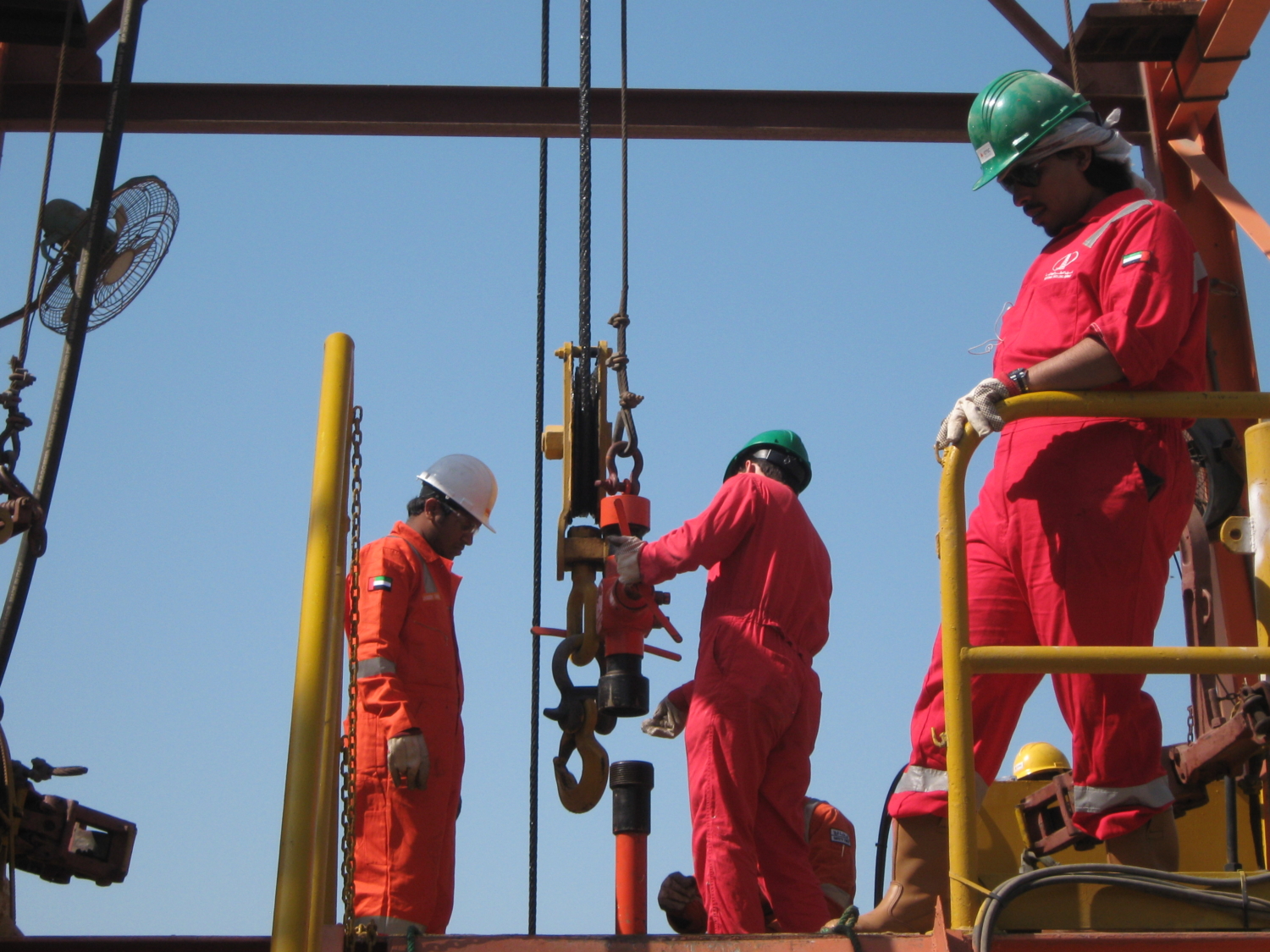 Workers in red coveralls and helmets operate equipment on an oil rig platform under a clear sky.