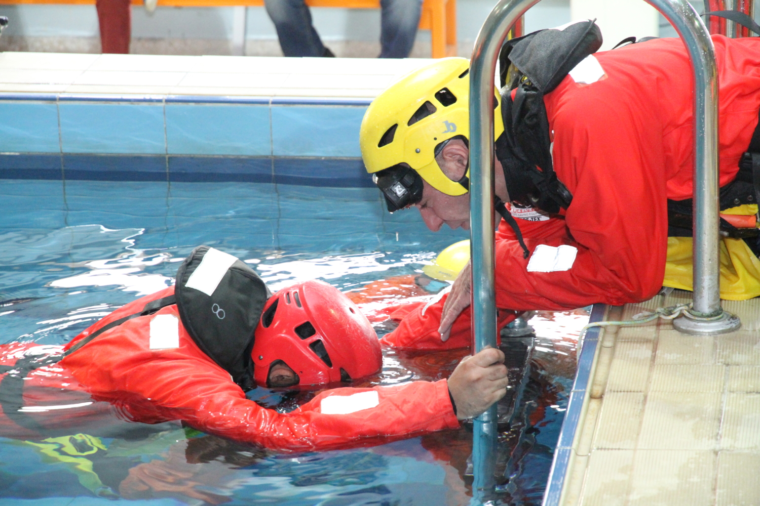 Two rescue workers in red suits and helmets perform a pool training exercise, with one standing in the water holding the pool edge and the other leaning over.