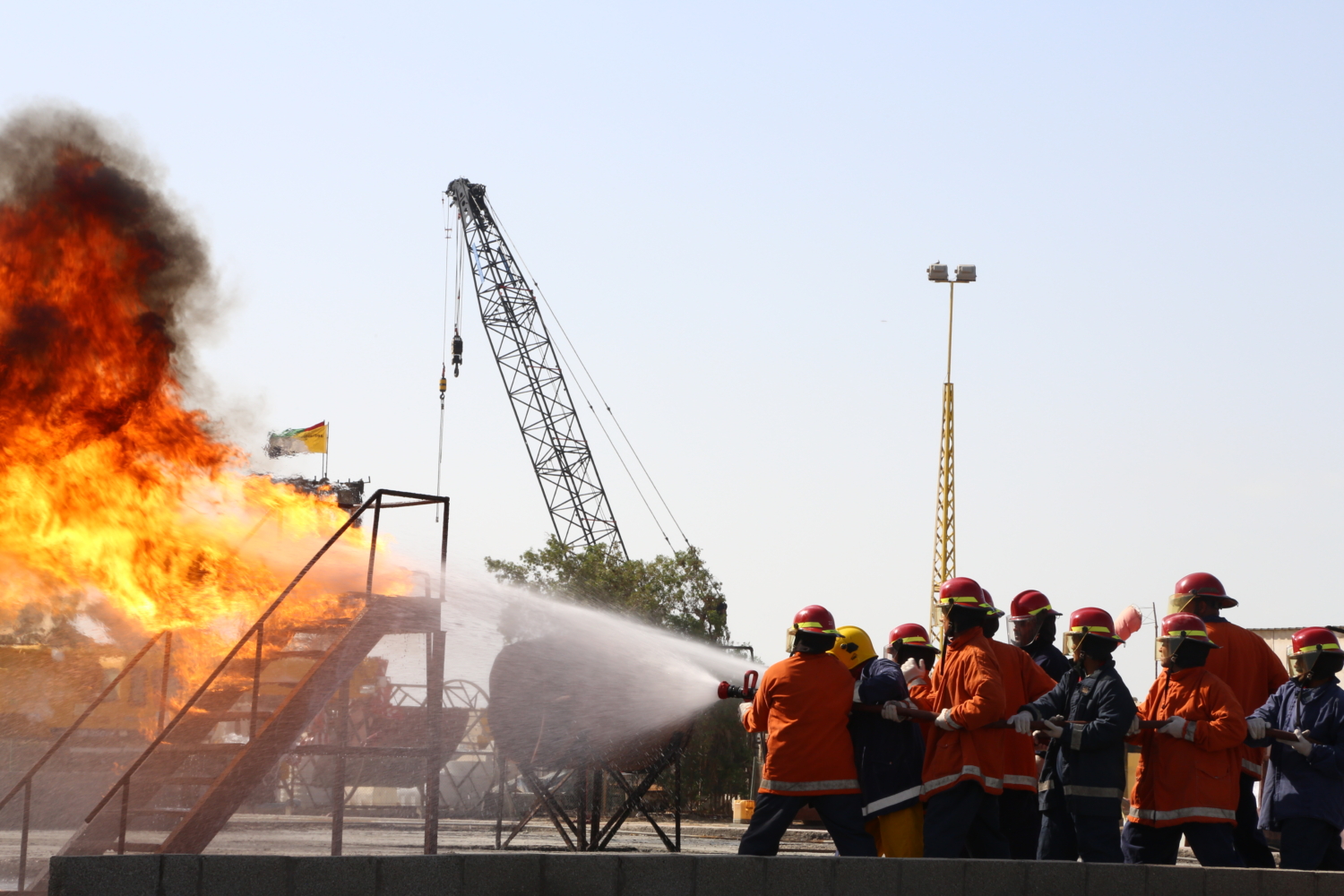 Firefighters in orange uniforms and helmets use a hose to extinguish a large fire during a daytime training exercise. A crane is visible in the background.