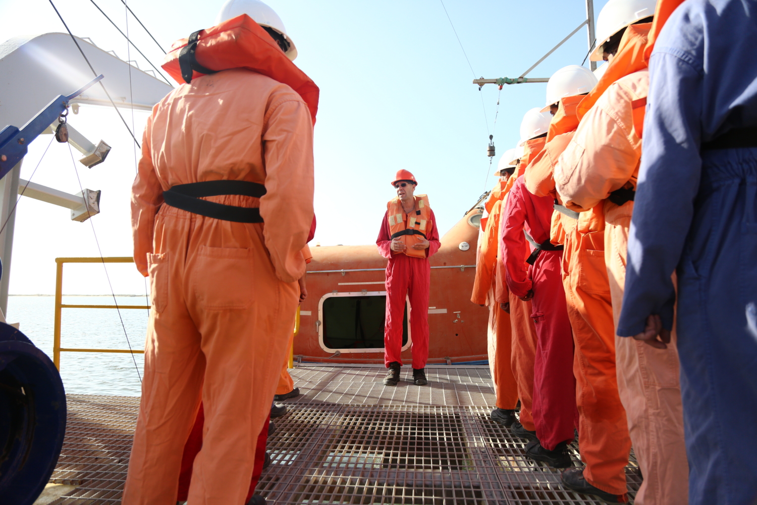 A group of people in safety helmets and colorful coveralls stand on a metal platform, facing a person in orange giving instructions.