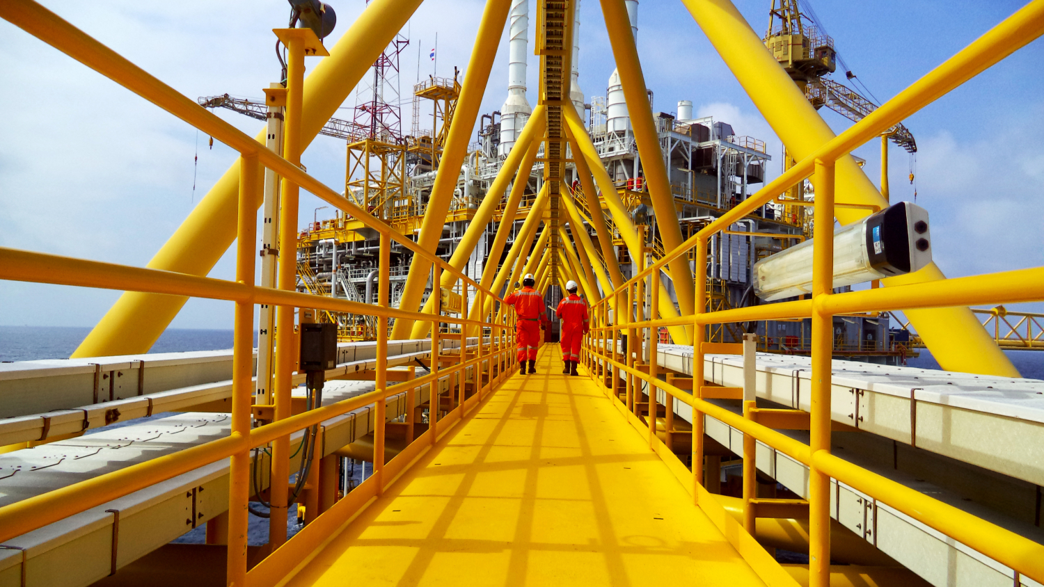 yellow walkway with two people in orange overalls walking towards an oil and gas rig