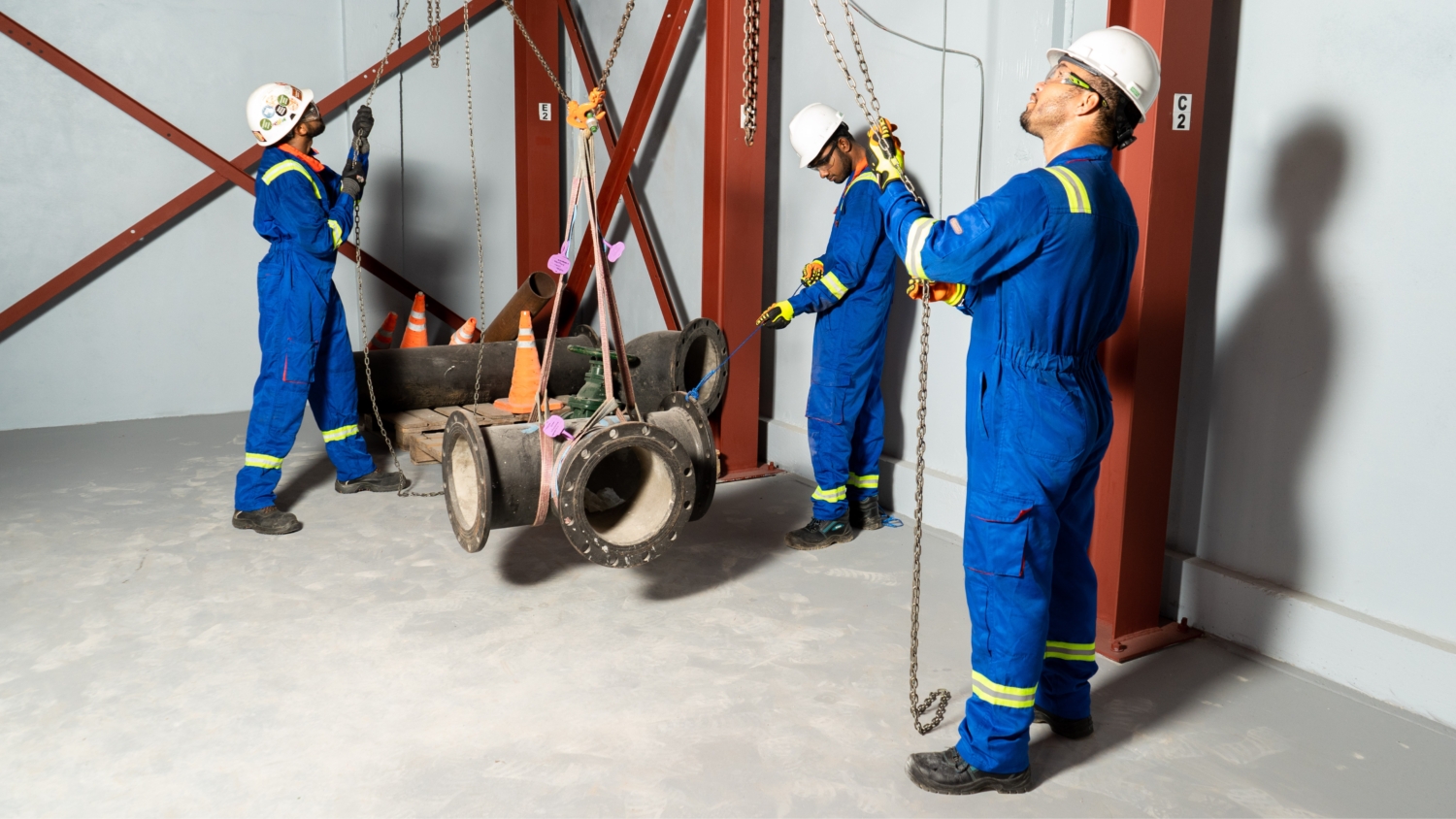 3 men in blue overalls using pulleys and chain to lift heavy pipe parts